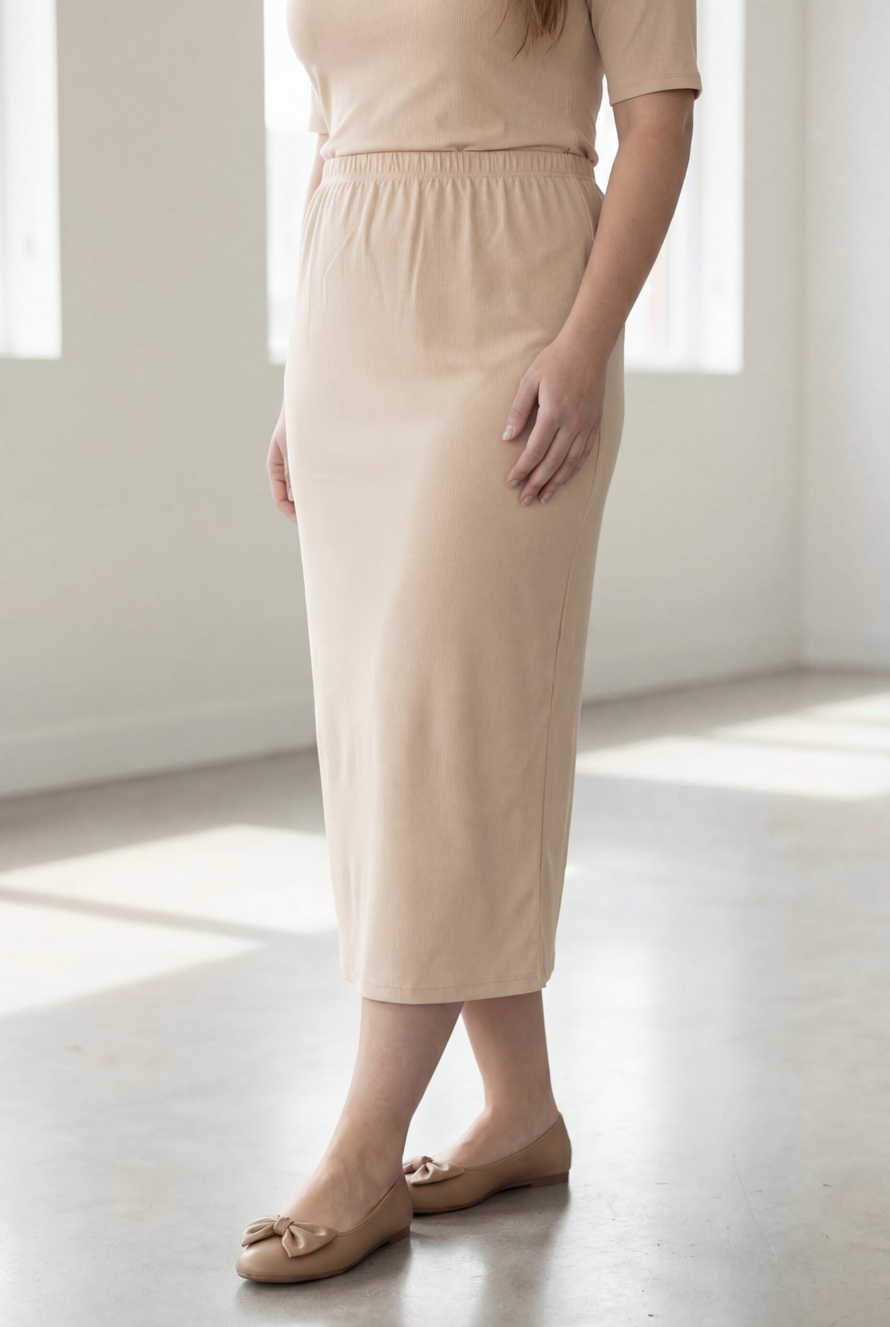 Woman wearing a beige shirt & maxi length pencil skirt standing in a bright room with large windows.