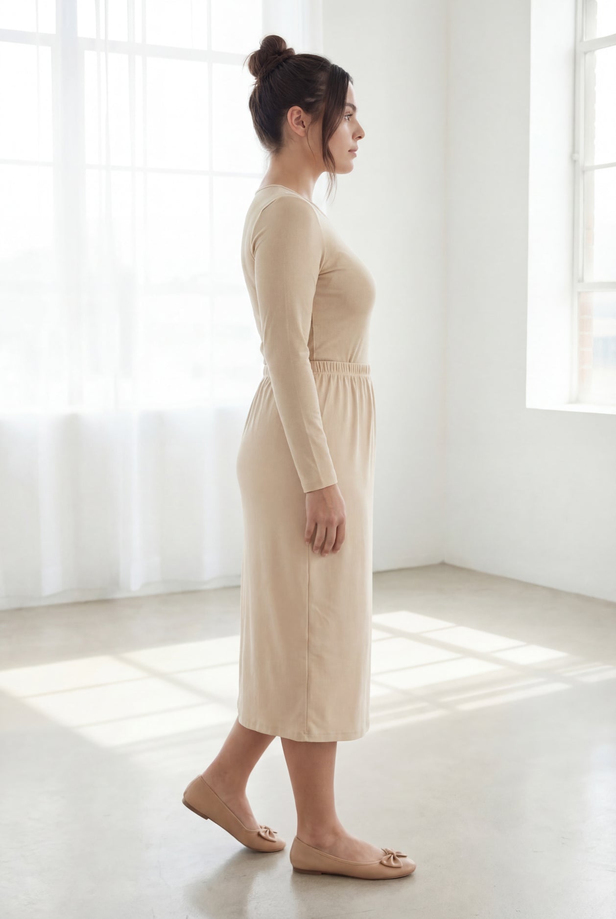 Woman wearing a beige shirt & skirt standing in a bright, minimalistic room.