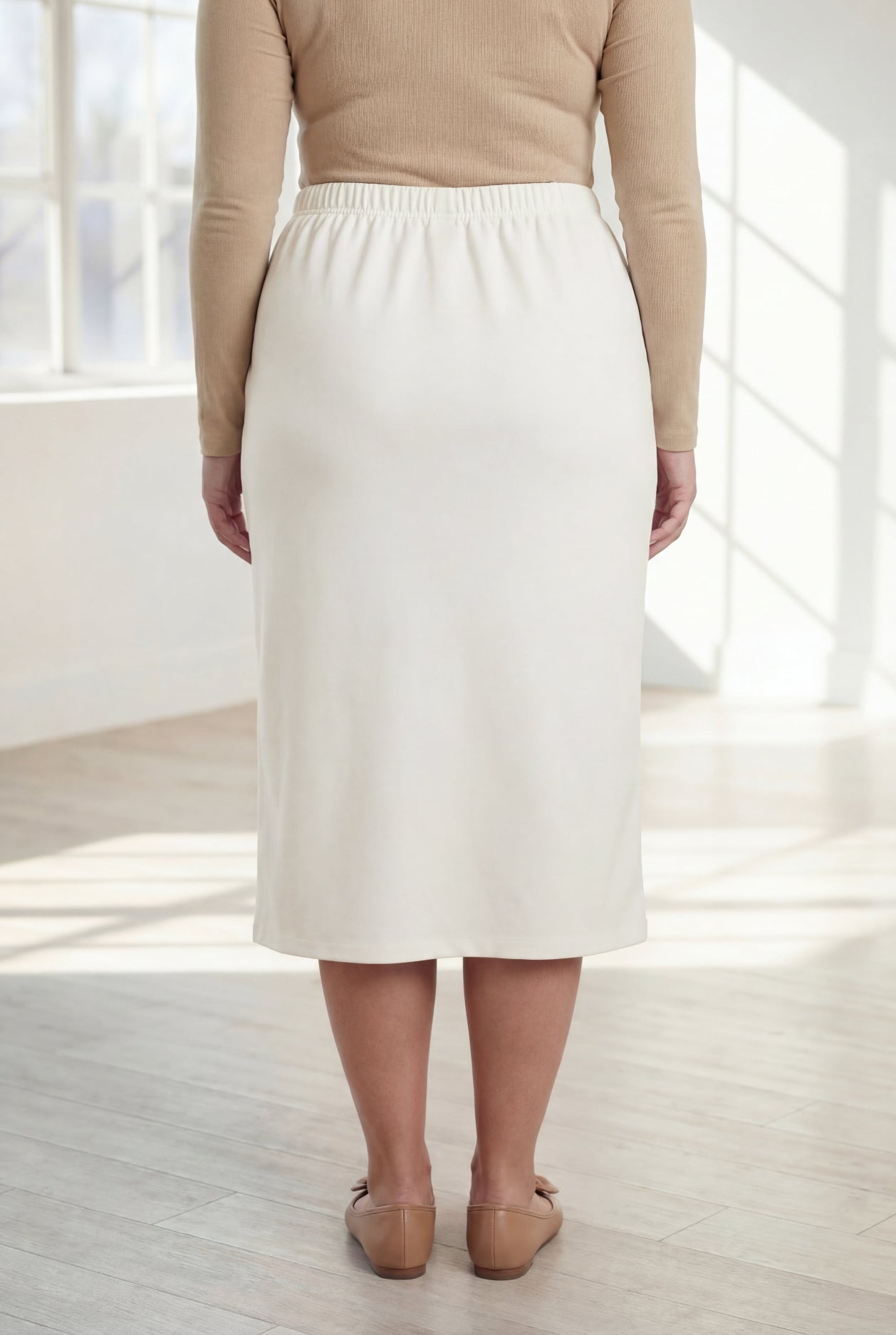 Woman wearing a beige top and white skirt in a bright room with large windows.