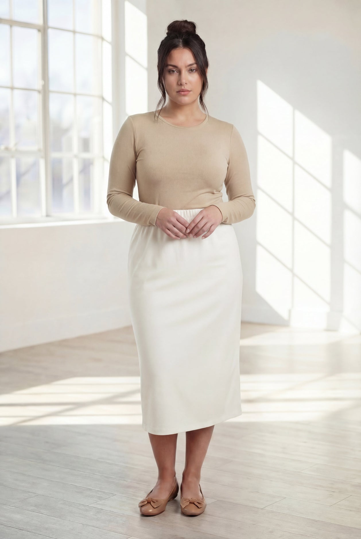 Woman wearing a beige top and white skirt in a bright room with large windows.