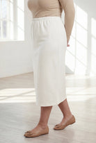 Woman wearing a beige top and white skirt in a bright room with large windows.