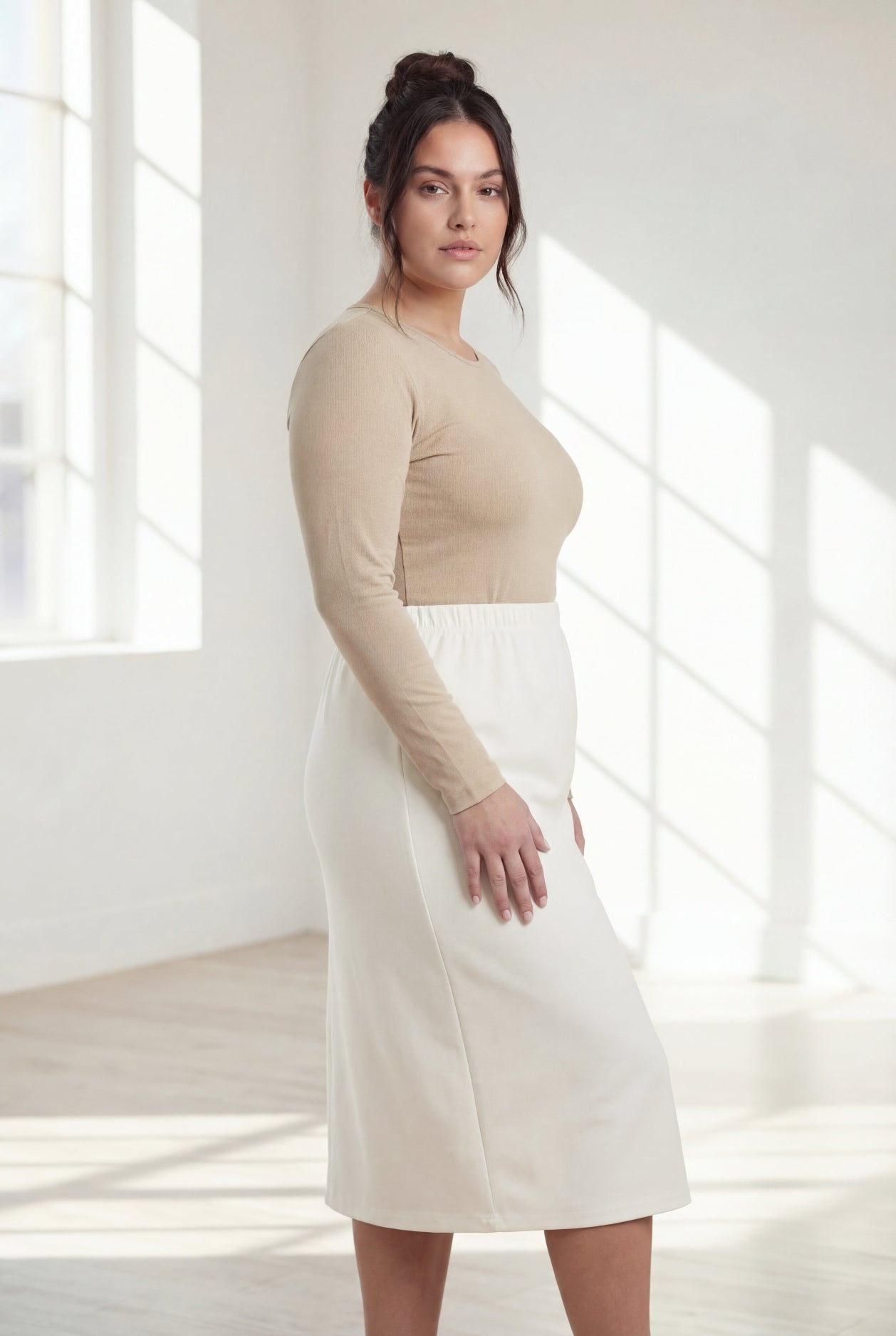 Woman wearing a beige top and white skirt in a bright room with large windows.