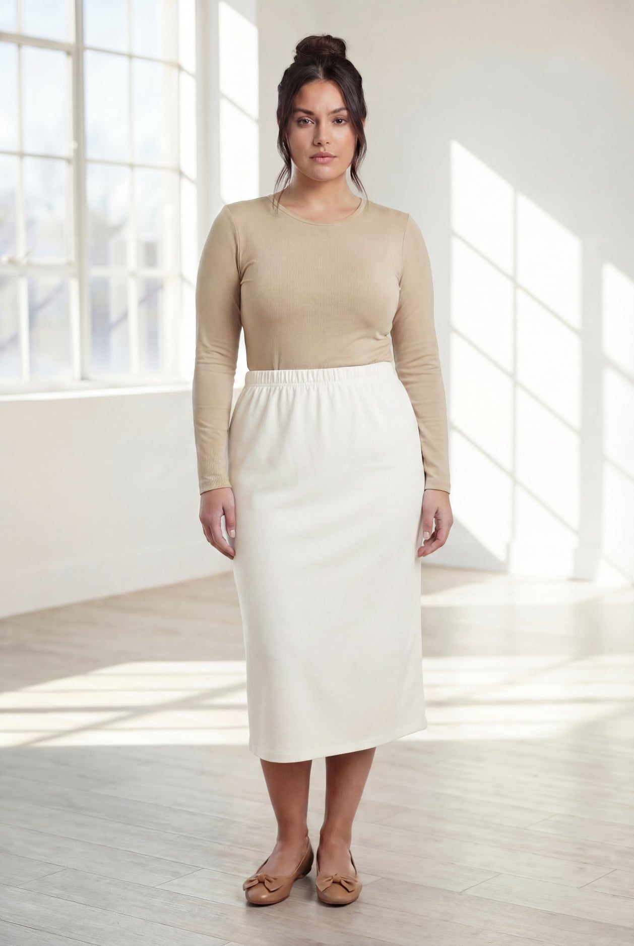 Woman wearing a beige top and white skirt in a bright room with large windows.