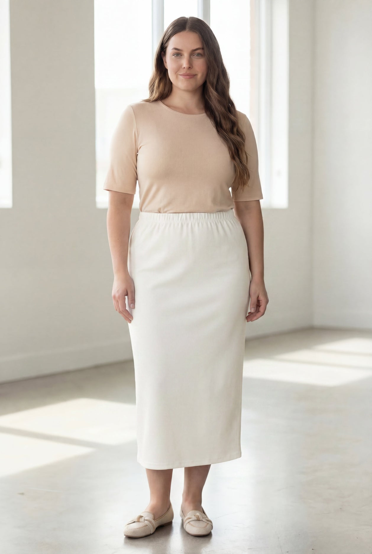 Woman wearing a beige shirt & off white maxi length pencil skirt standing in a bright room with large windows.