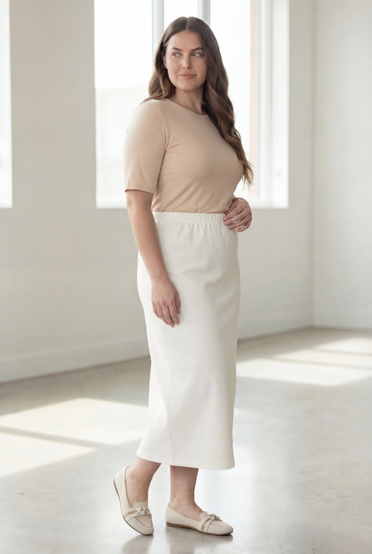 Woman wearing a beige shirt & off white maxi length pencil skirt standing in a bright room with large windows.