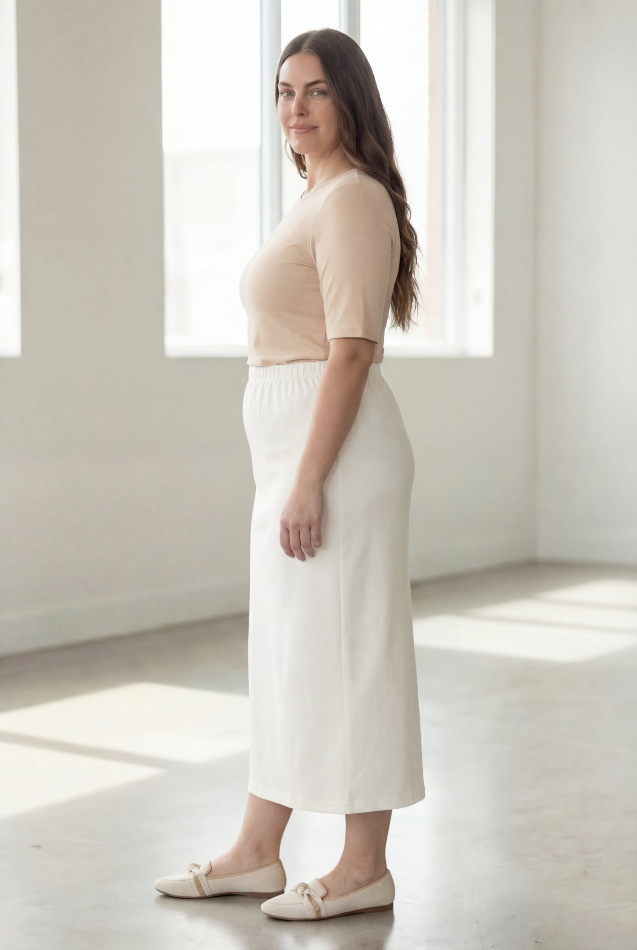 Woman wearing a beige shirt & off white maxi length pencil skirt standing in a bright room with large windows.