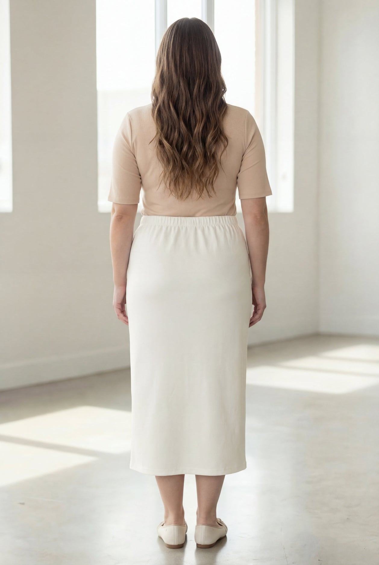 Woman wearing a beige shirt & off white maxi length pencil skirt standing in a bright room with large windows.