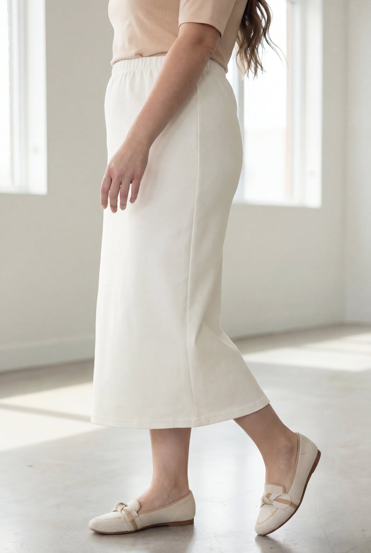 Woman wearing a beige shirt & off white maxi length pencil skirt standing in a bright room with large windows.