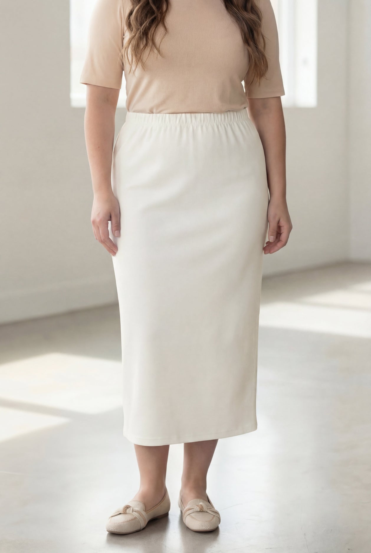 Woman wearing a beige shirt & off white maxi length pencil skirt standing in a bright room with large windows.
