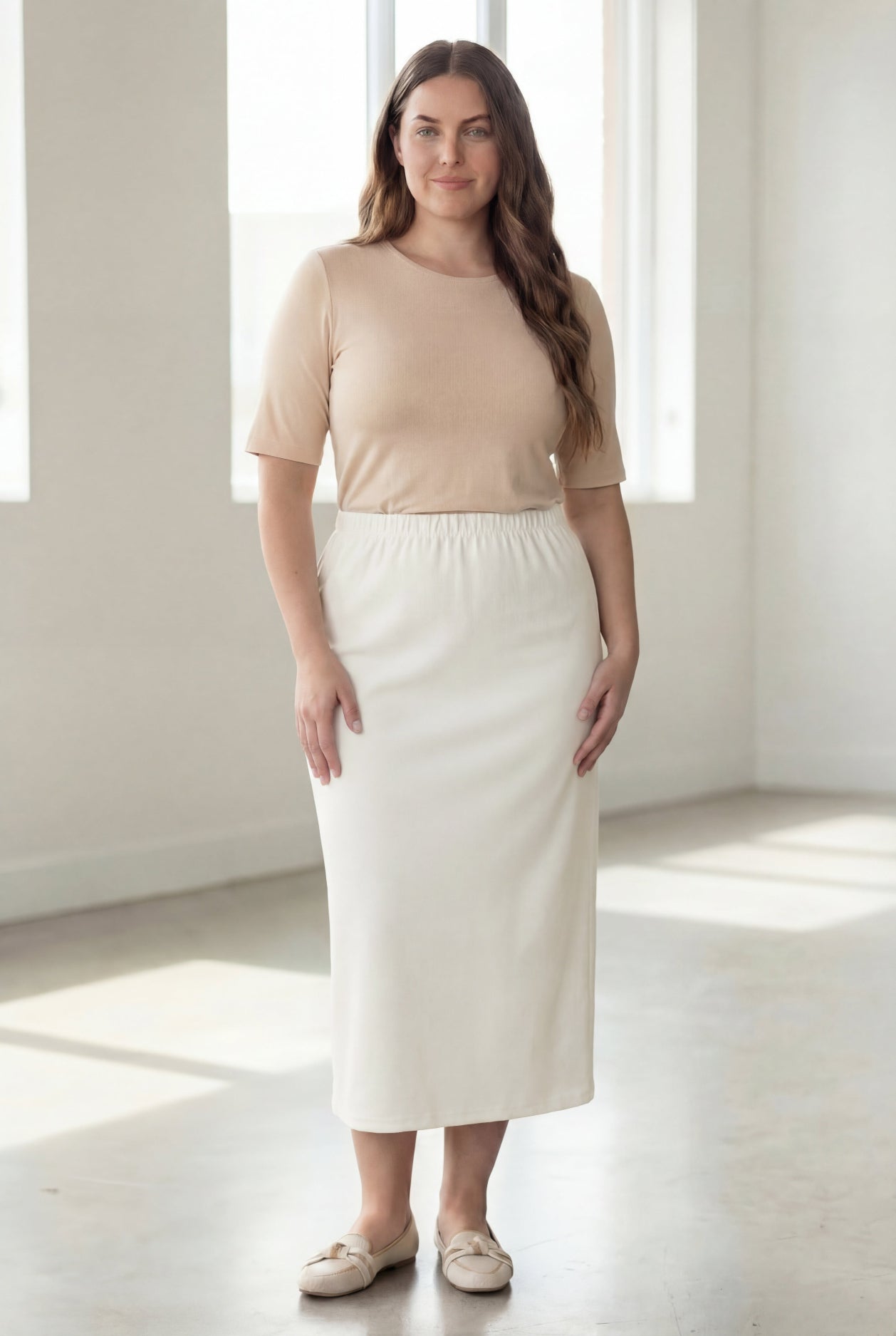 Woman wearing a beige shirt & off white maxi length pencil skirt standing in a bright room with large windows.