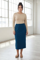 Woman wearing a beige top and blue skirt in a bright room with large windows.