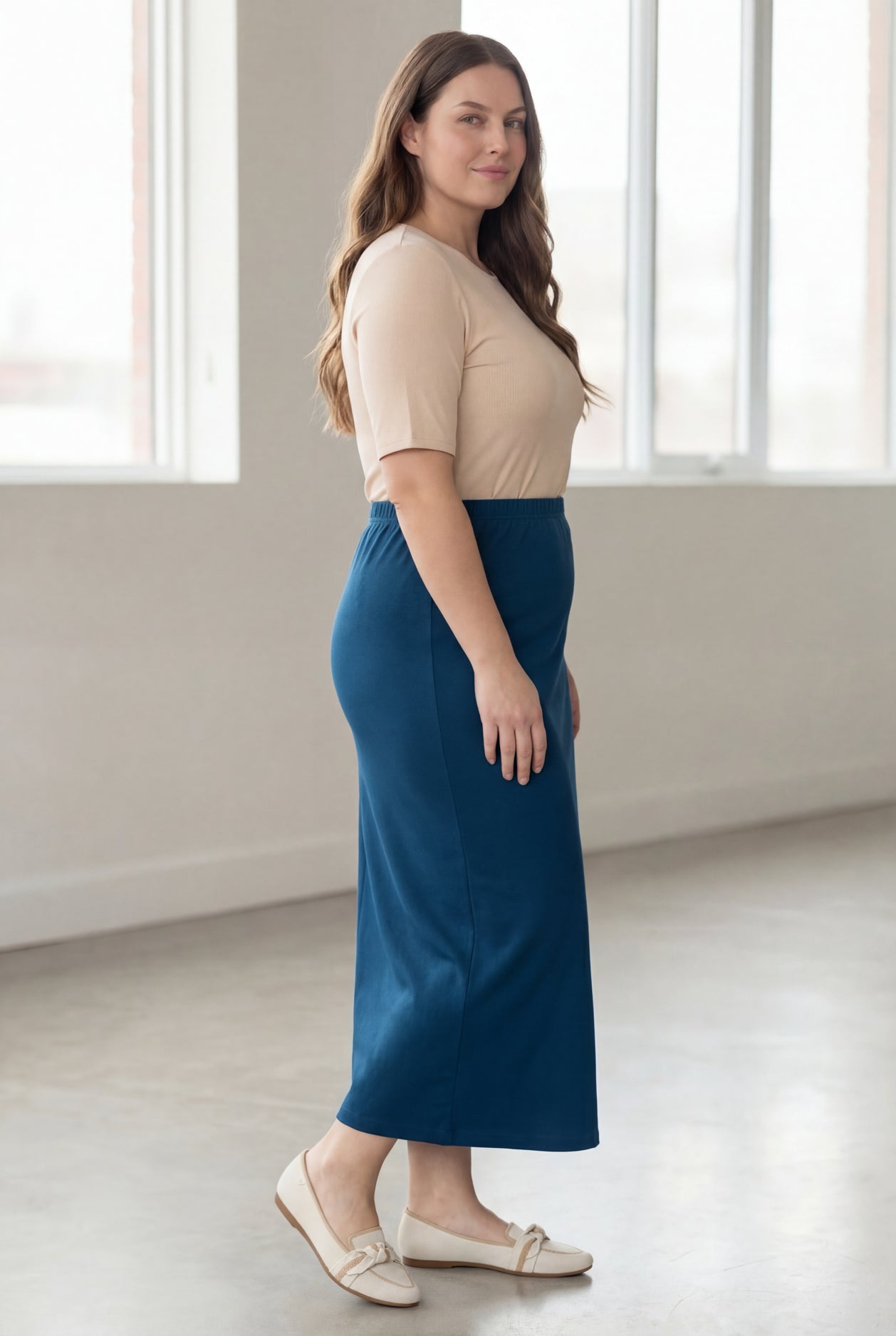 Woman wearing a beige shirt & blue maxi length pencil skirt standing in a bright room with large windows.