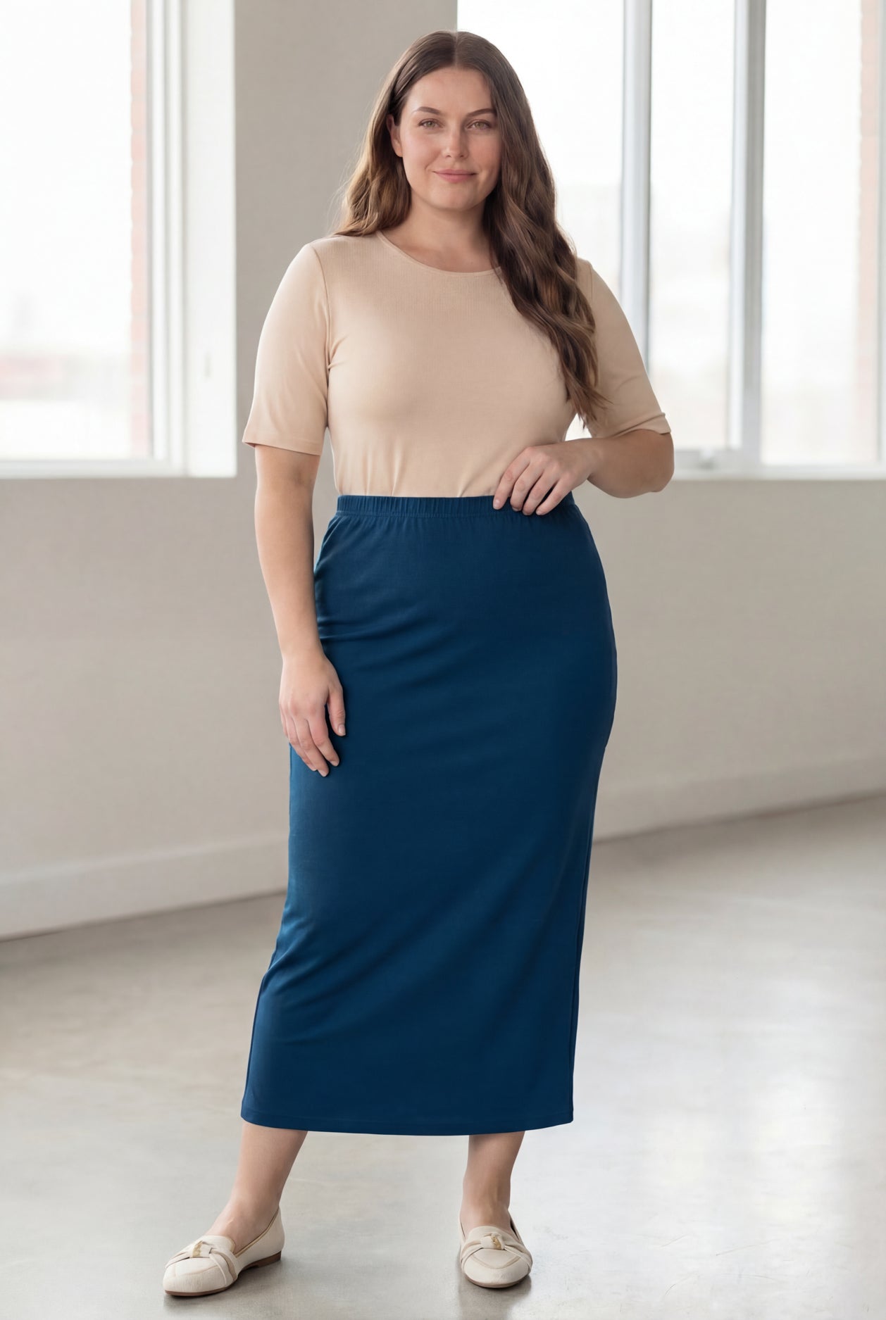 Woman wearing a beige shirt & blue maxi length pencil skirt standing in a bright room with large windows.