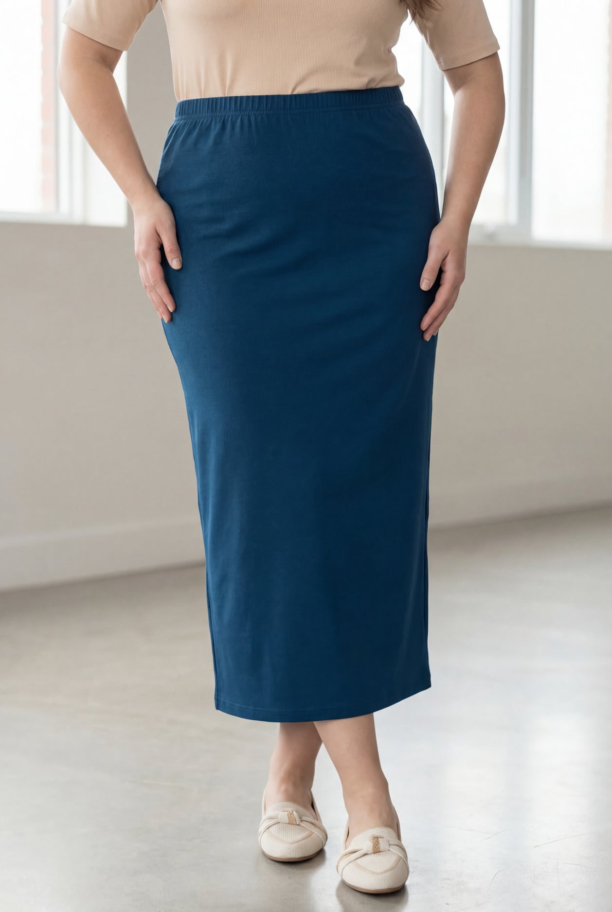 Woman wearing a beige shirt & blue maxi length pencil skirt standing in a bright room with large windows.
