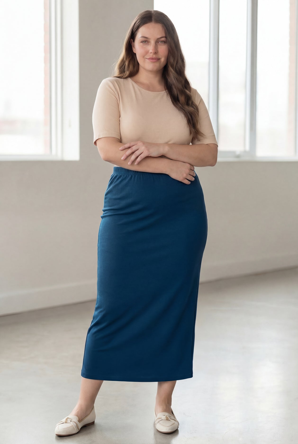 Woman wearing a beige shirt & blue maxi length pencil skirt standing in a bright room with large windows.