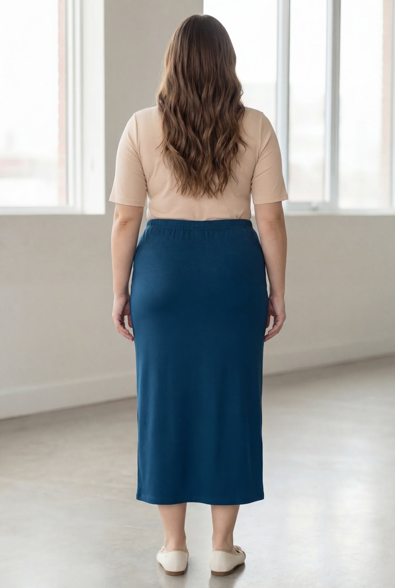 Woman wearing a beige shirt & blue maxi length pencil skirt standing in a bright room with large windows.