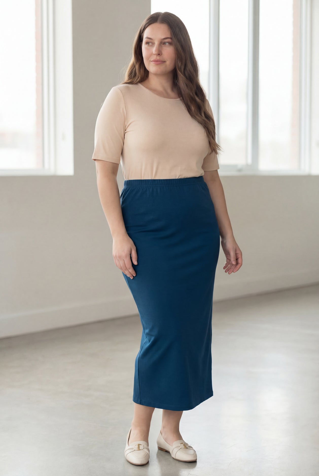 Woman wearing a beige shirt & blue maxi length pencil skirt standing in a bright room with large windows.