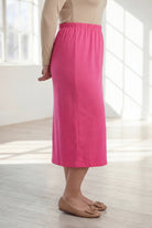 Woman wearing a beige top and pink skirt in a bright room with large windows.