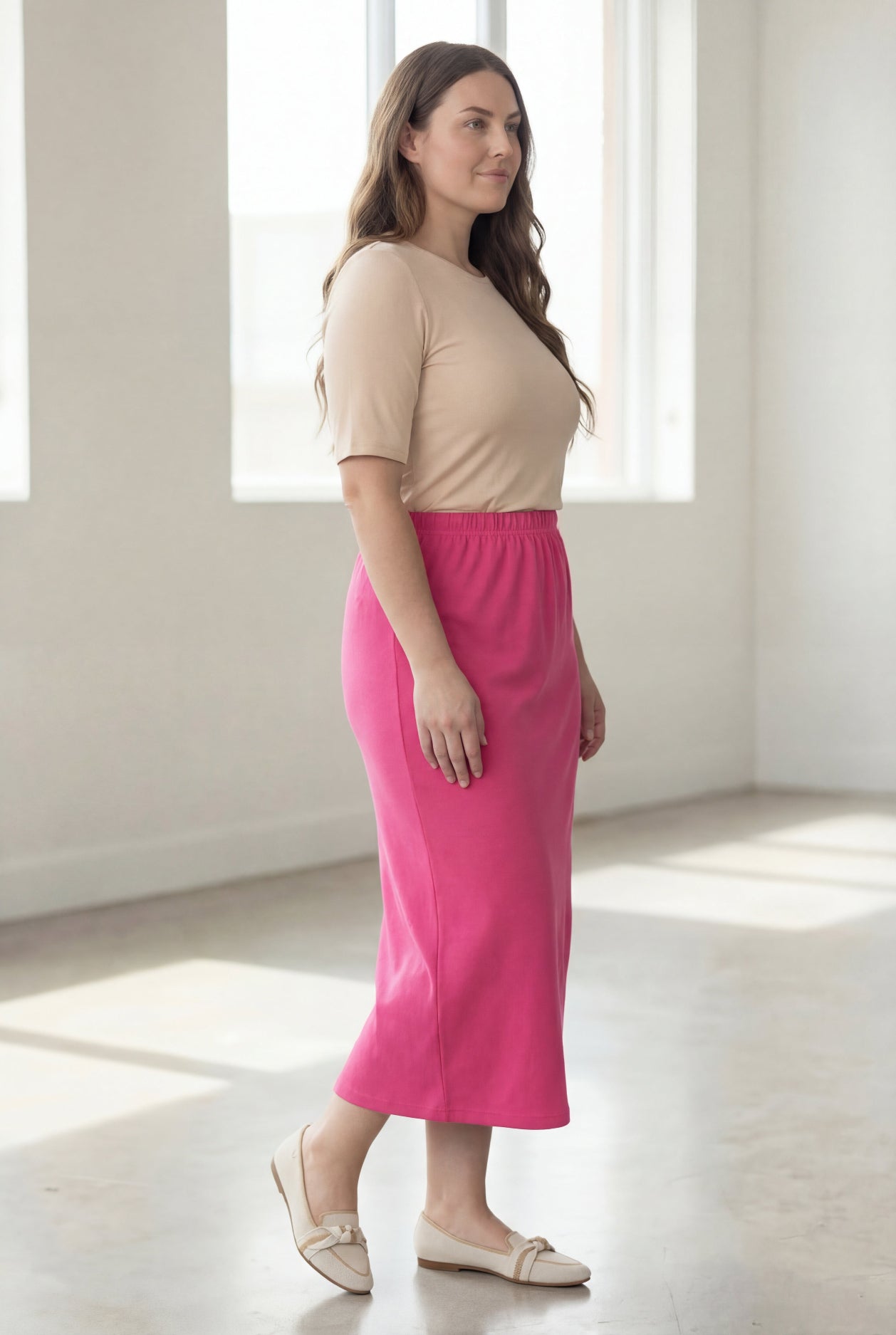 Woman wearing a beige shirt & pink maxi length pencil skirt standing in a bright room with large windows.