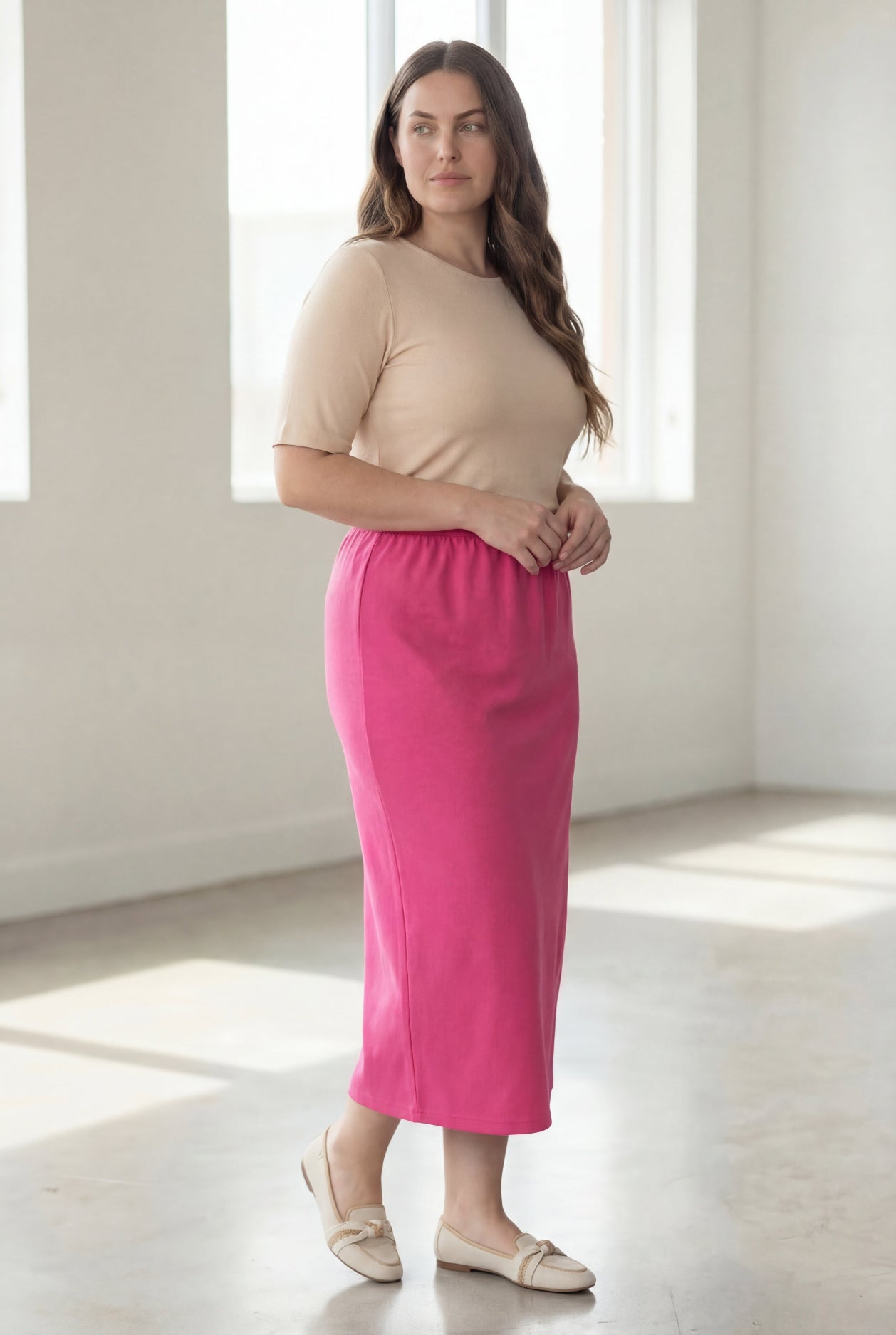 Woman wearing a beige shirt & pink maxi length pencil skirt standing in a bright room with large windows.