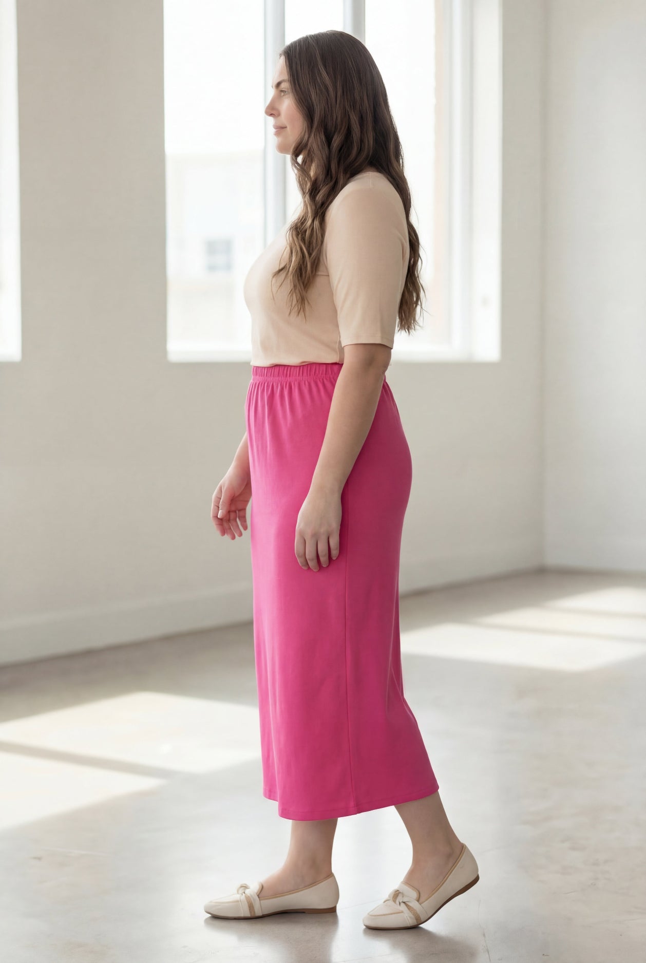 Woman wearing a beige shirt & pink maxi length pencil skirt standing in a bright room with large windows.