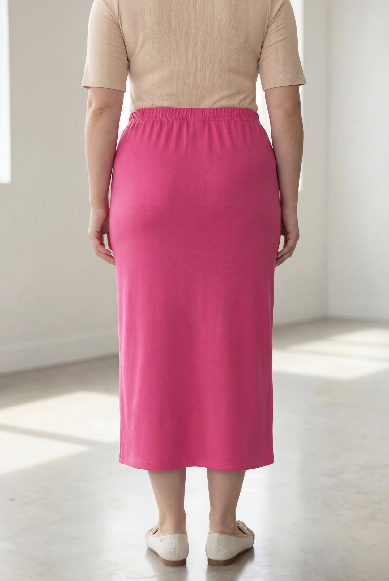 Woman wearing a beige shirt & pink maxi length pencil skirt standing in a bright room with large windows.