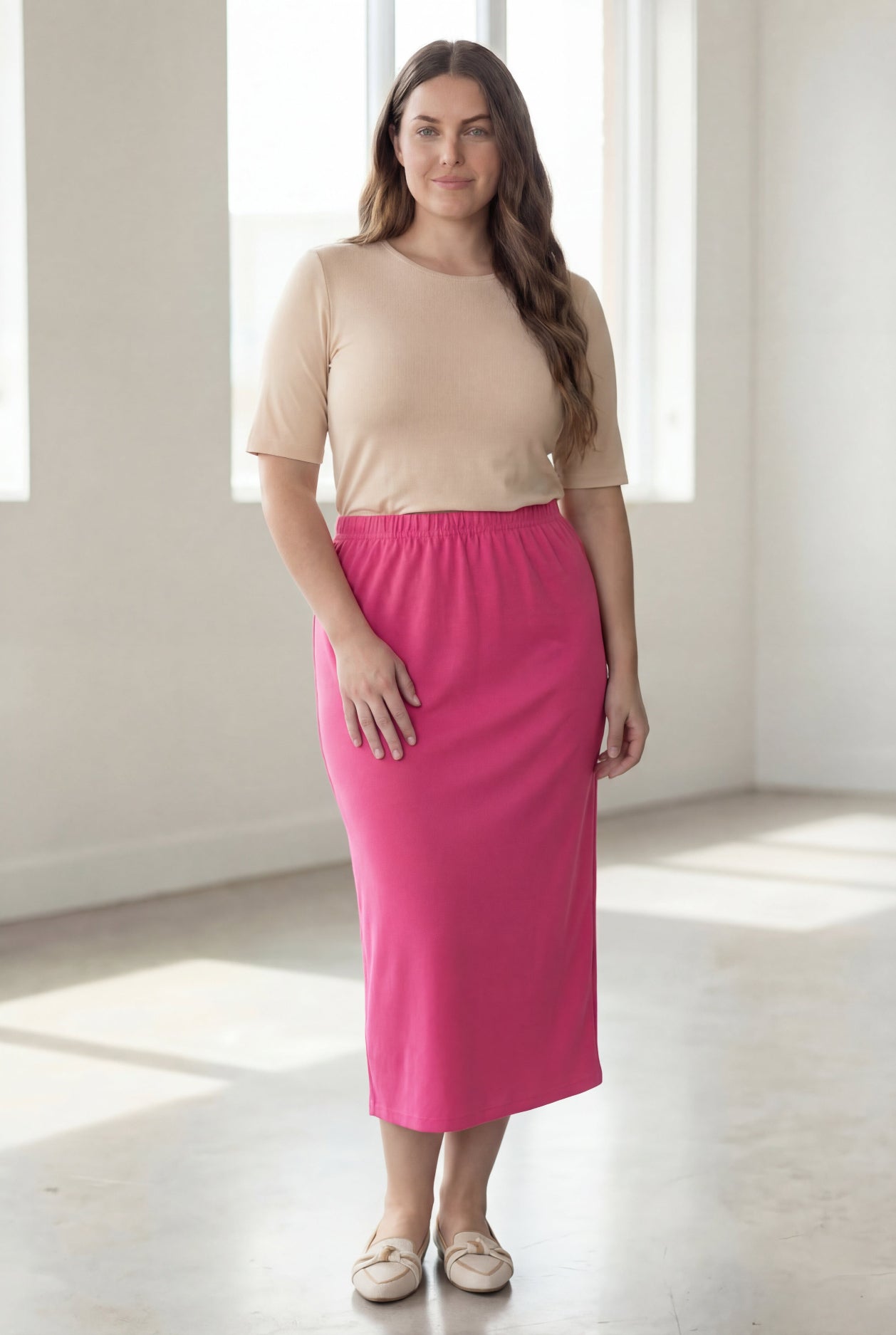 Woman wearing a beige shirt & pink maxi length pencil skirt standing in a bright room with large windows.