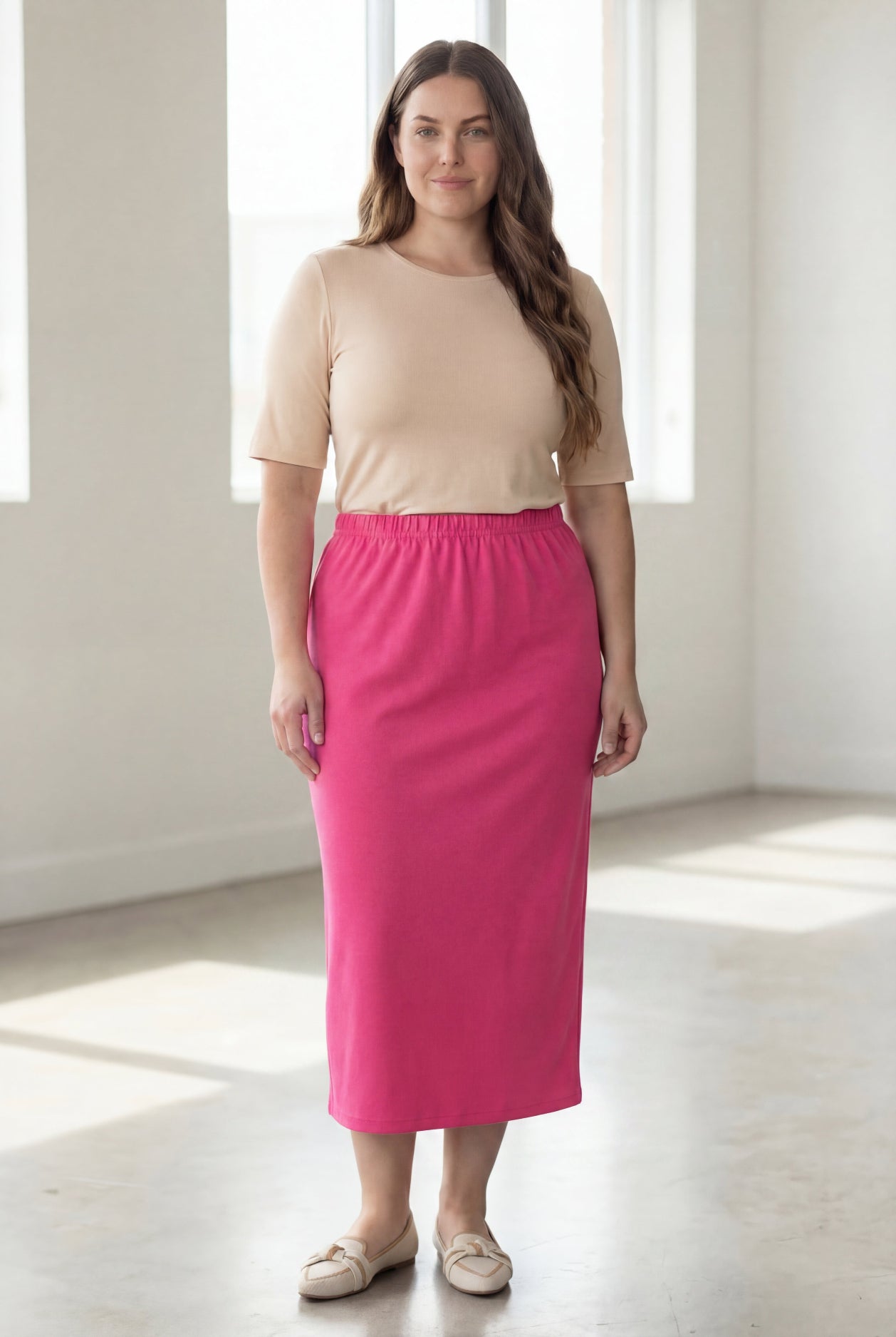 Woman wearing a beige shirt & pink maxi length pencil skirt standing in a bright room with large windows.