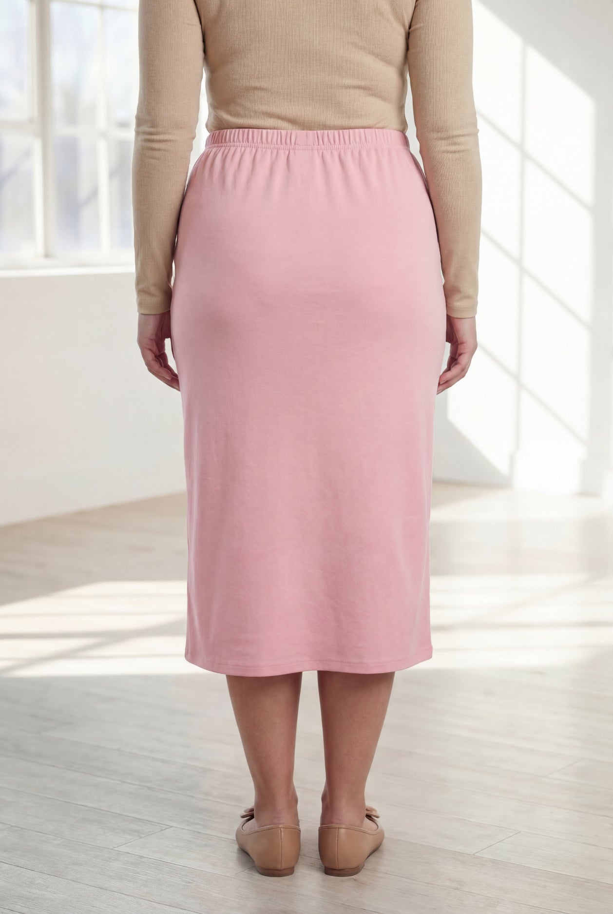Woman wearing a beige top and pink skirt in a bright room with large windows.