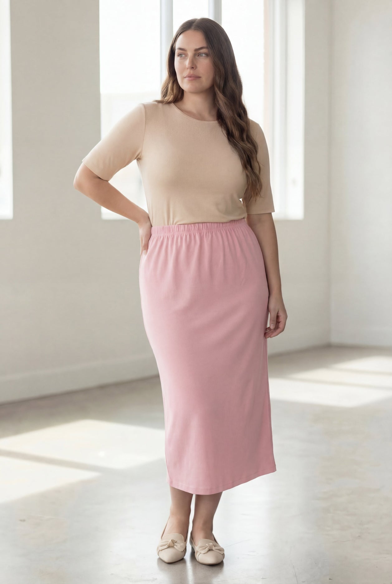 Woman wearing a beige shirt & pink maxi length pencil skirt standing in a bright room with large windows.