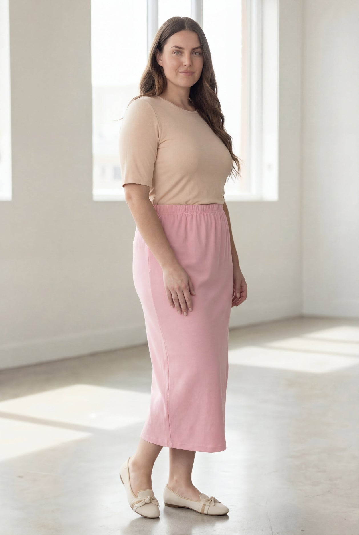 Woman wearing a beige shirt & pink maxi length pencil skirt standing in a bright room with large windows.