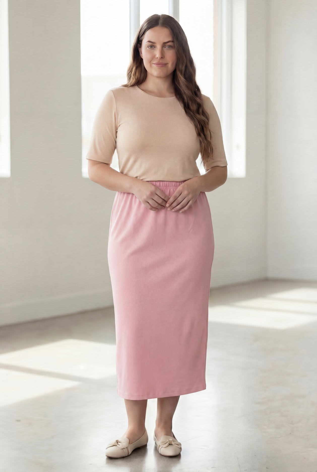 Woman wearing a beige shirt & pink maxi length pencil skirt standing in a bright room with large windows.