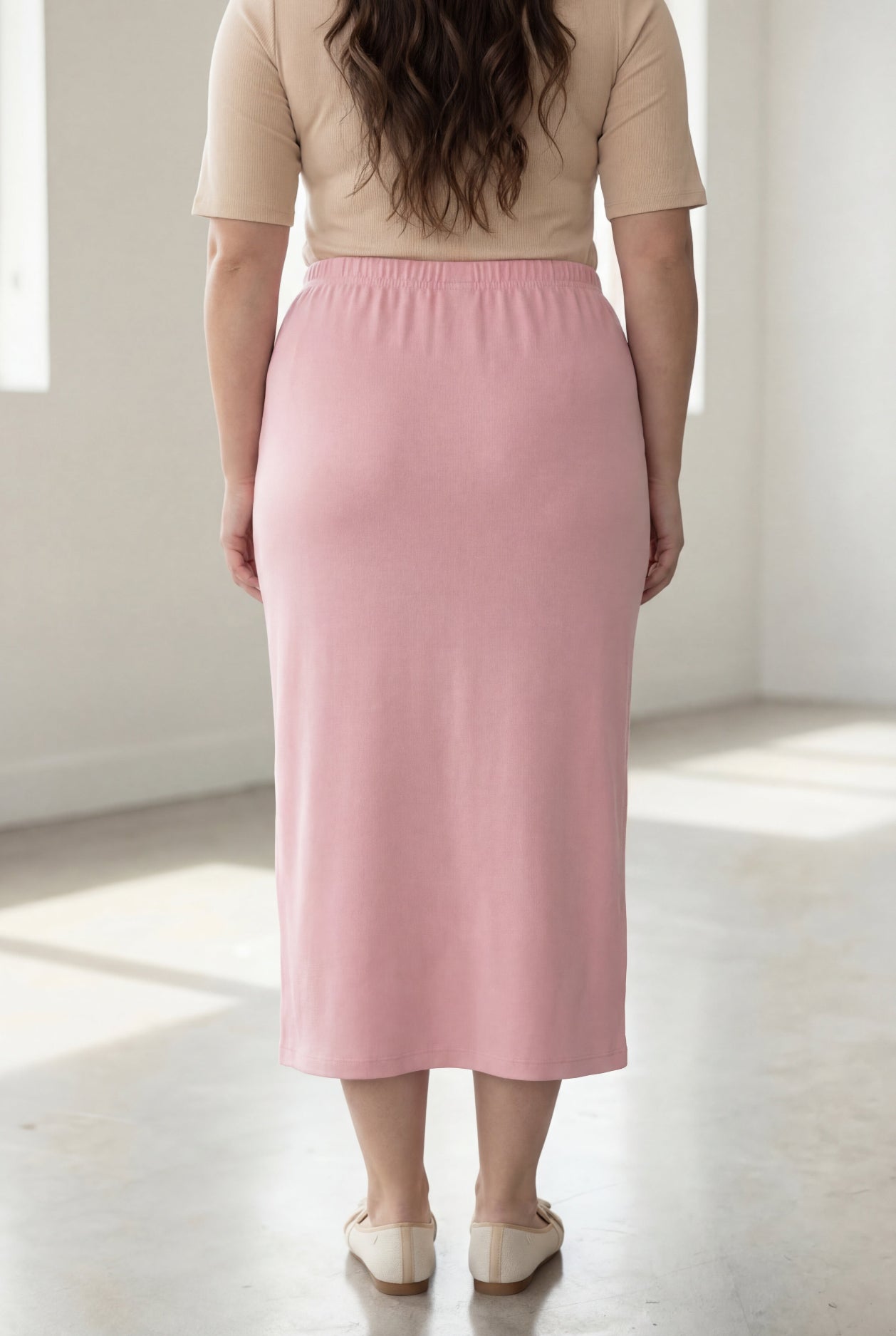 Woman wearing a beige shirt & pink maxi length pencil skirt standing in a bright room with large windows.
