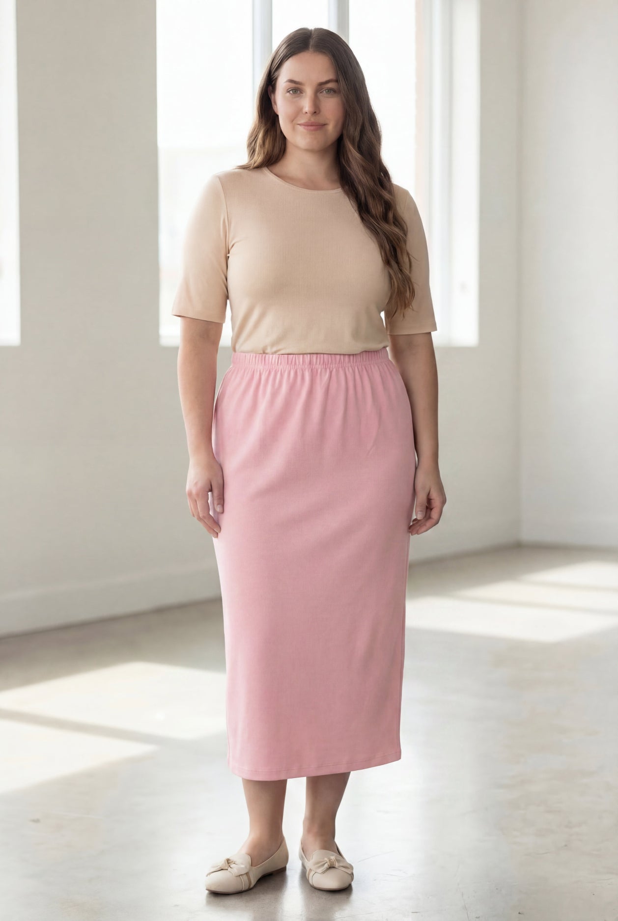 Woman wearing a beige shirt & pink maxi length pencil skirt standing in a bright room with large windows.