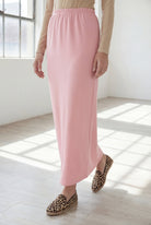 Woman wearing a beige shirt & pink maxi length pencil skirt standing in a bright room with large windows.