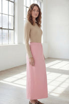Woman wearing a beige shirt & pink maxi length pencil skirt standing in a bright room with large windows.