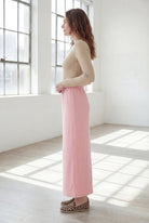 Woman wearing a beige shirt & pink maxi length pencil skirt standing in a bright room with large windows.