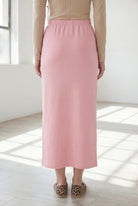 Woman wearing a beige shirt & pink maxi length pencil skirt standing in a bright room with large windows.