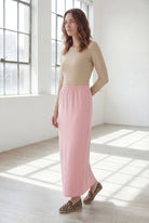 Woman wearing a beige shirt & pink maxi length pencil skirt standing in a bright room with large windows.