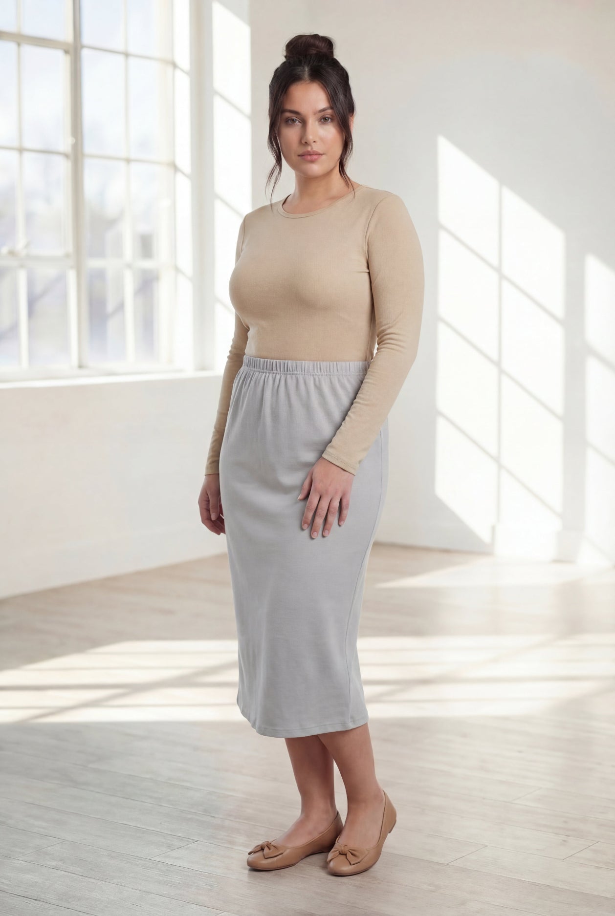 Woman wearing a beige top and silver skirt in a bright room with large windows.
