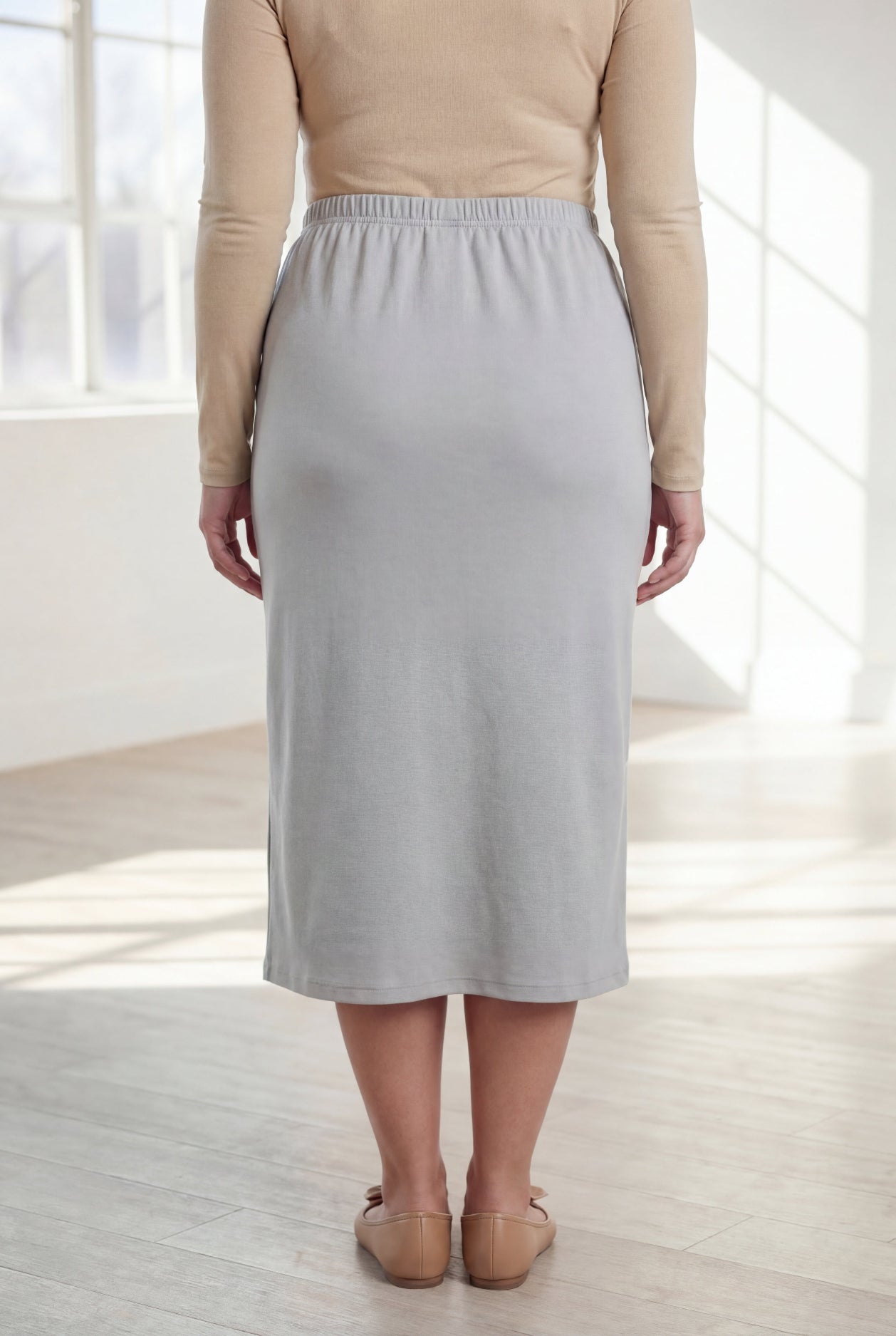 Woman wearing a beige top and silver skirt in a bright room with large windows.