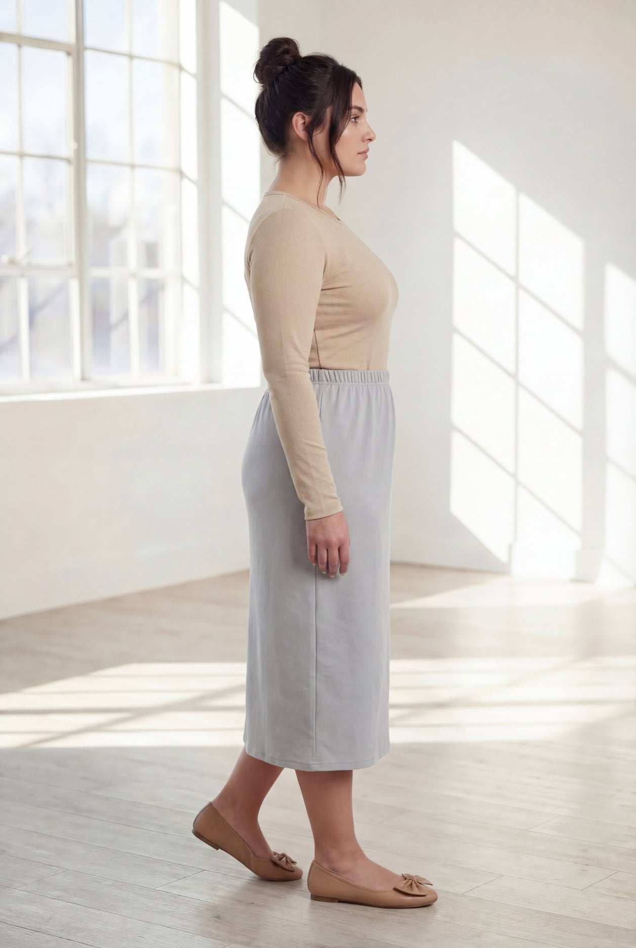 Woman wearing a beige top and silver skirt in a bright room with large windows.