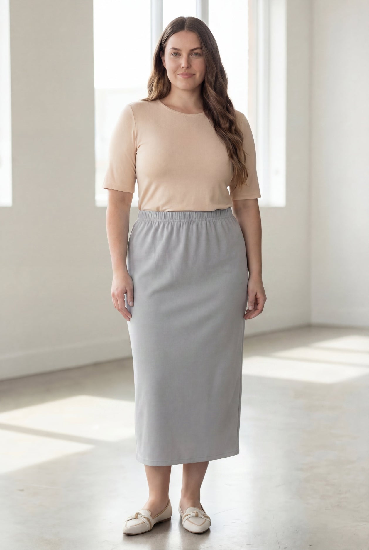 Woman wearing a beige shirt & silver maxi length pencil skirt standing in a bright room with large windows.