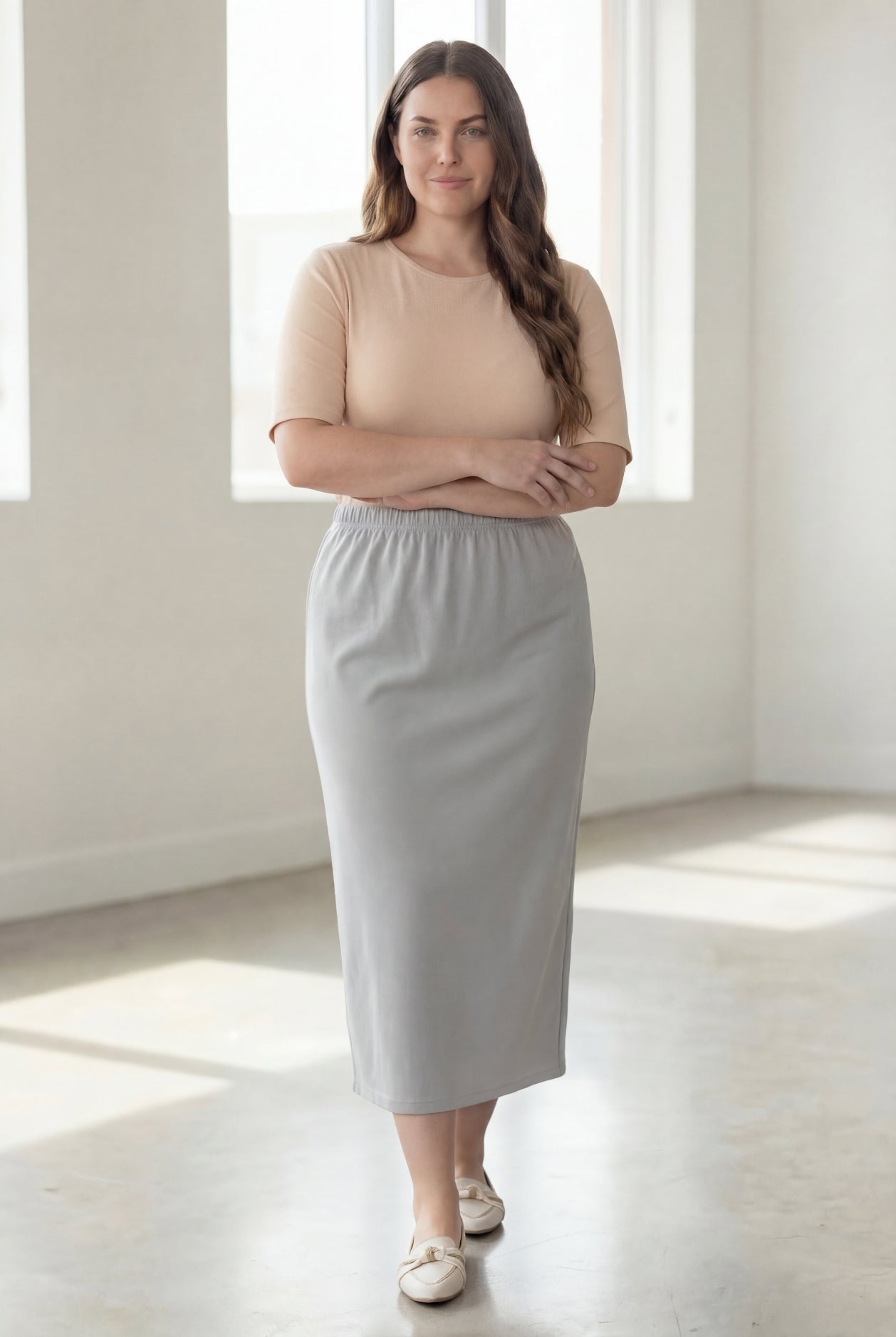 Woman wearing a beige shirt & silver maxi length pencil skirt standing in a bright room with large windows.