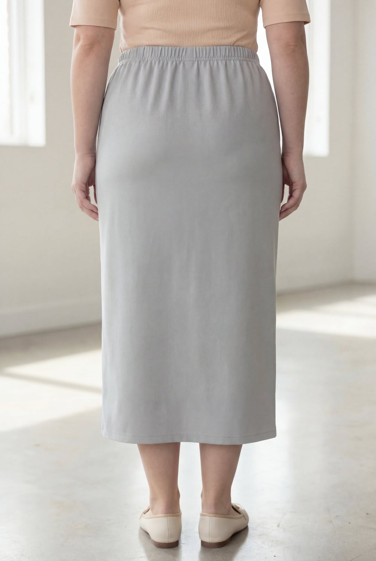 Woman wearing a beige shirt & silver maxi length pencil skirt standing in a bright room with large windows.