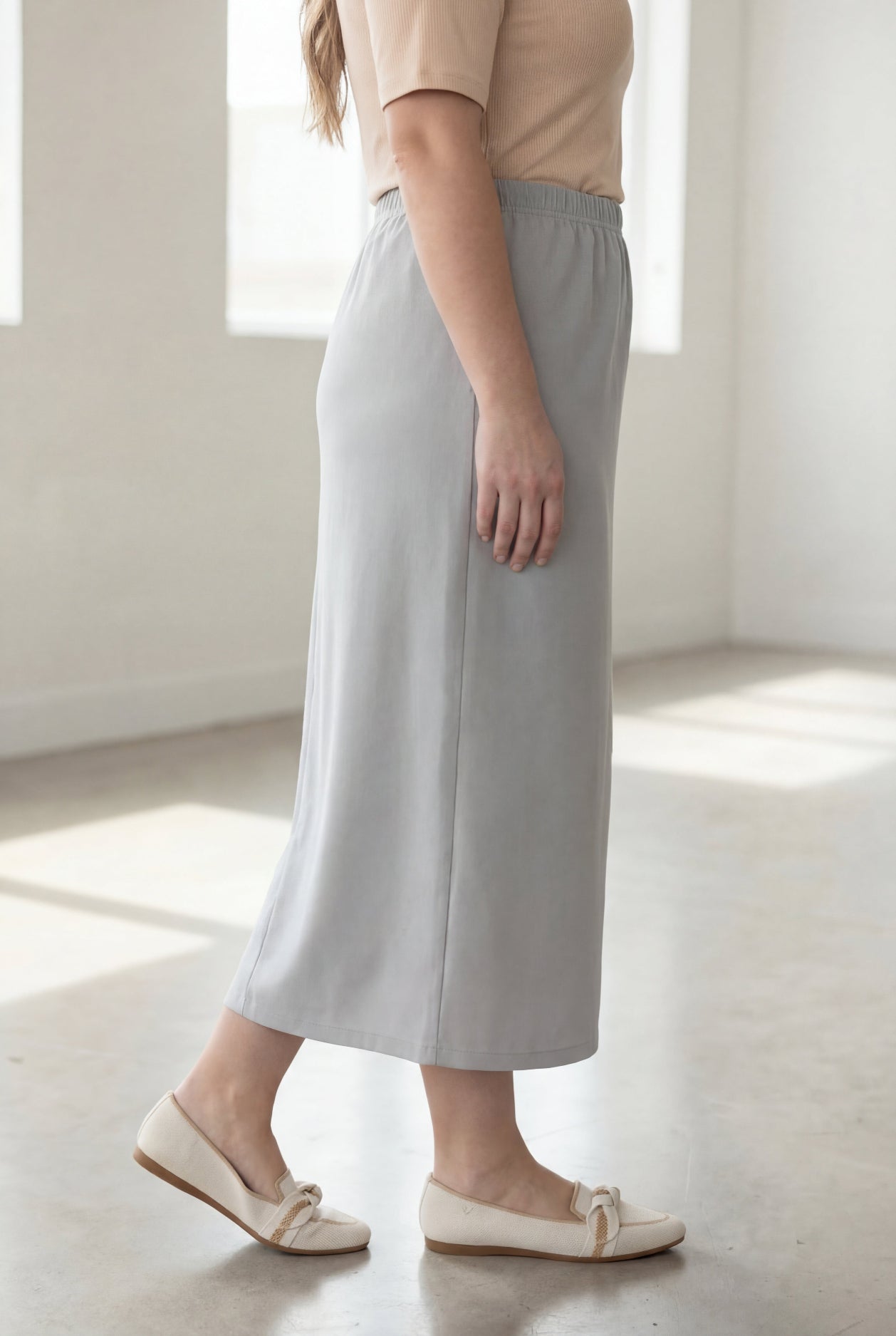 Woman wearing a beige shirt & silver maxi length pencil skirt standing in a bright room with large windows.