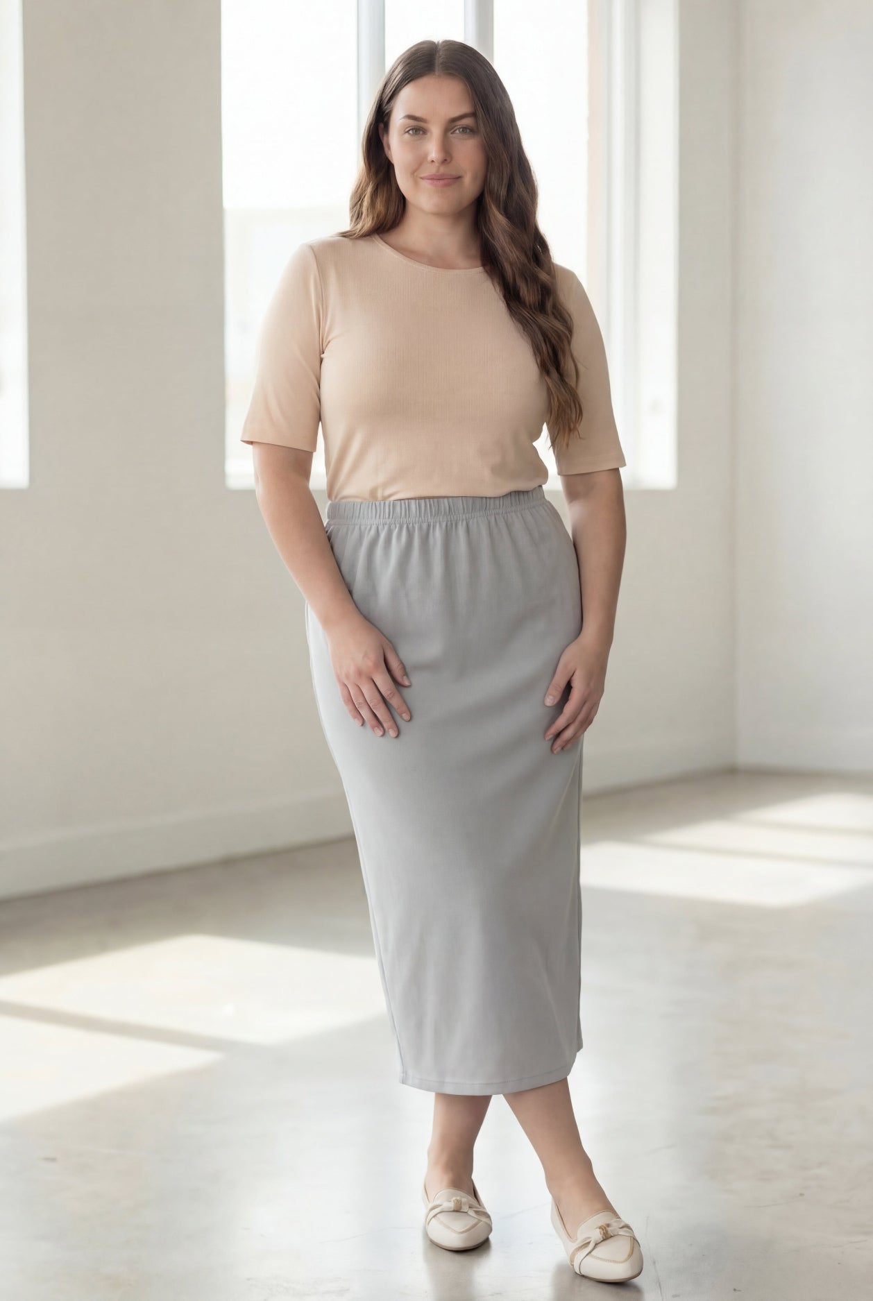 Woman wearing a beige shirt & silver maxi length pencil skirt standing in a bright room with large windows.