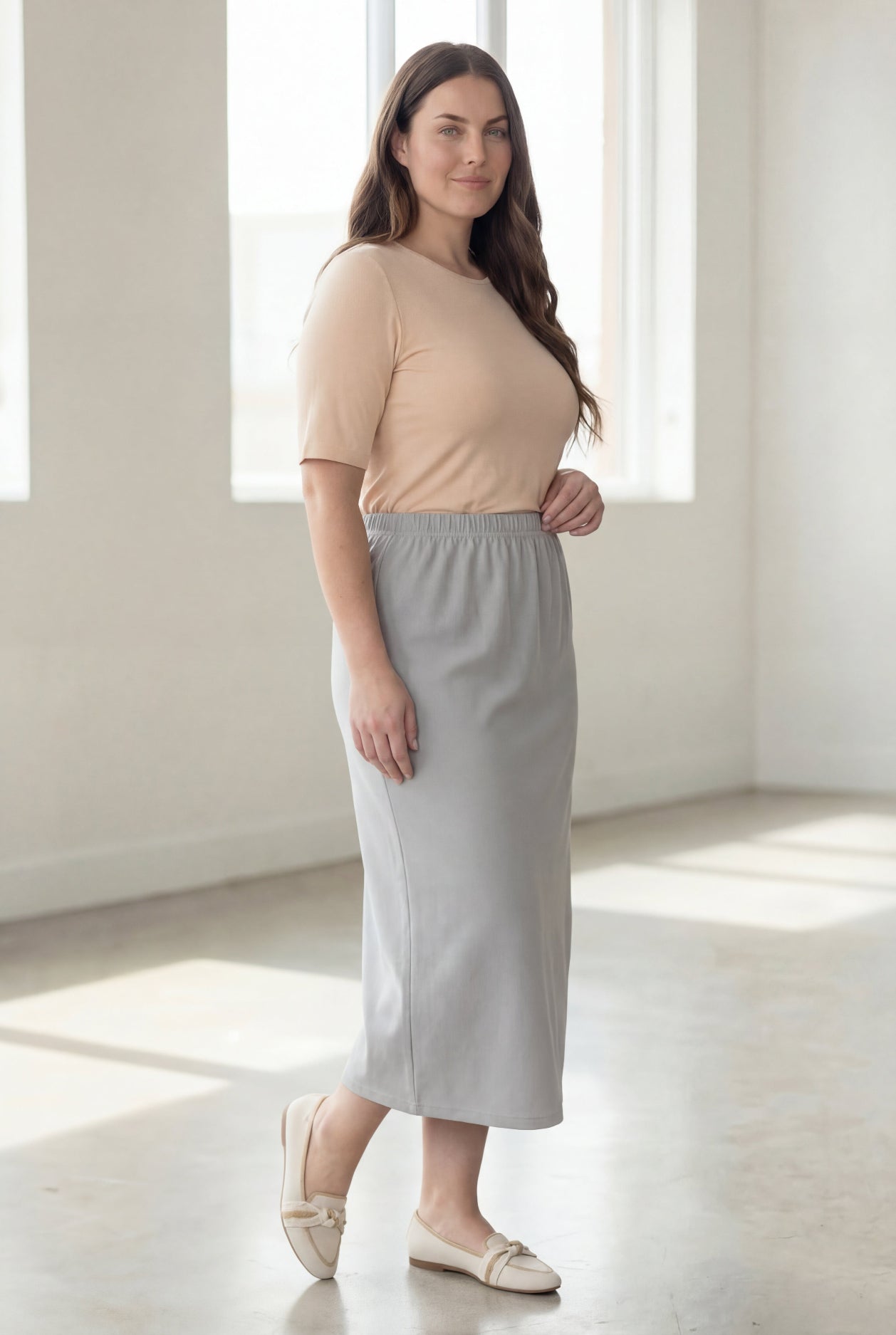 Woman wearing a beige shirt & silver maxi length pencil skirt standing in a bright room with large windows.